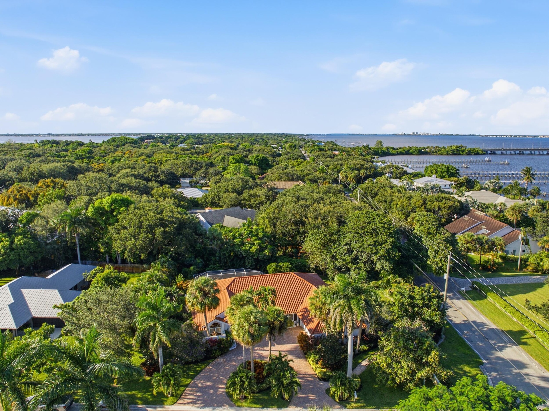 Aerial perspective of a single-story residence featuring a clay tile roof, paved circular driveway, mature landscaping with palm trees, and proximity 