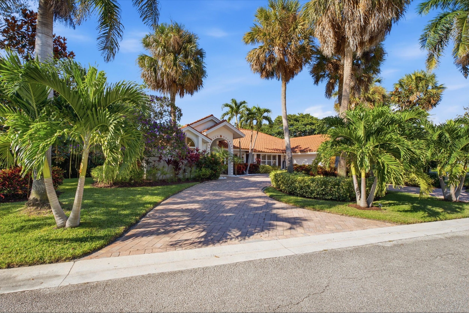 Exterior facade featuring a pitched tile roof, arched entryway, and brick-paver driveway surrounded by mature palm trees and manicured landscaping