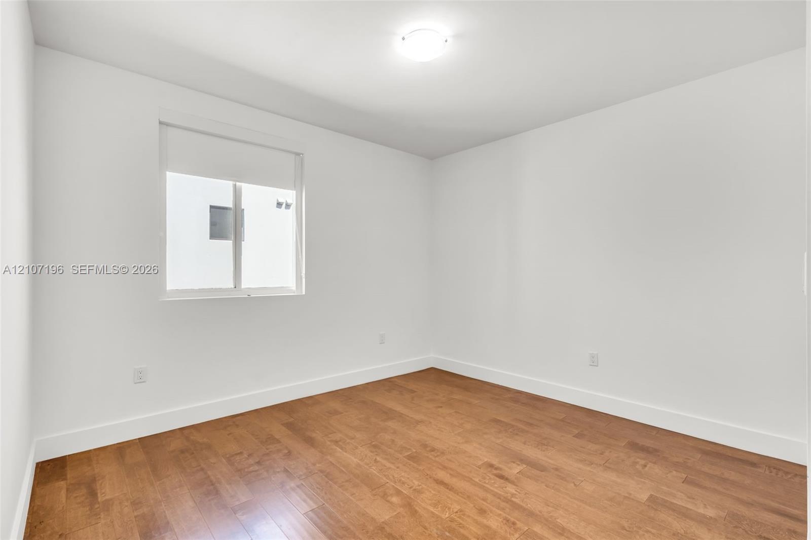 Room featuring wood-finish flooring, white walls, and a single-pane window with a roller shade, illuminated by a flush-mount ceiling light