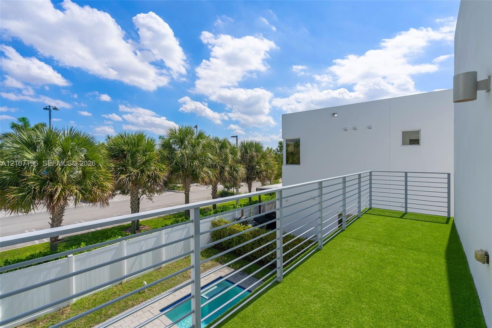 Primary Bedroom balcony featuring synthetic turf flooring, horizontal metal railings, exterior wall-mounted lighting, and smooth white stucco siding