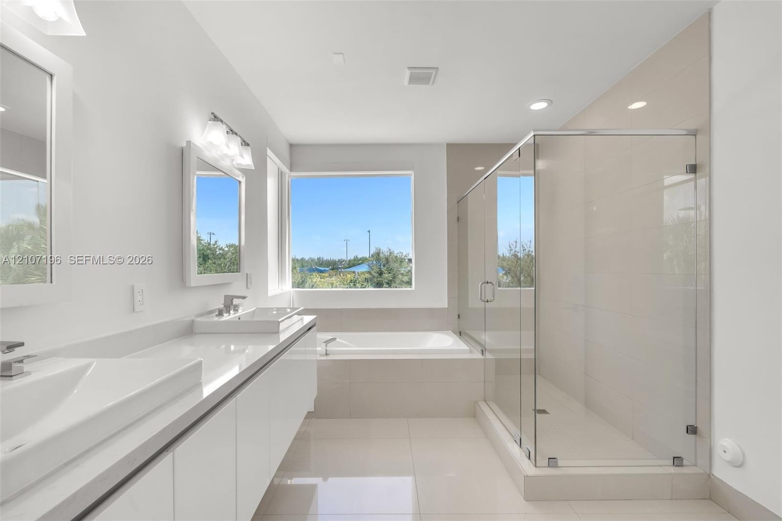 Dual-sink vanity with clean-lined cabinetry, built-in soaking tub, glass-enclosed shower, polished tile flooring, and large picture window