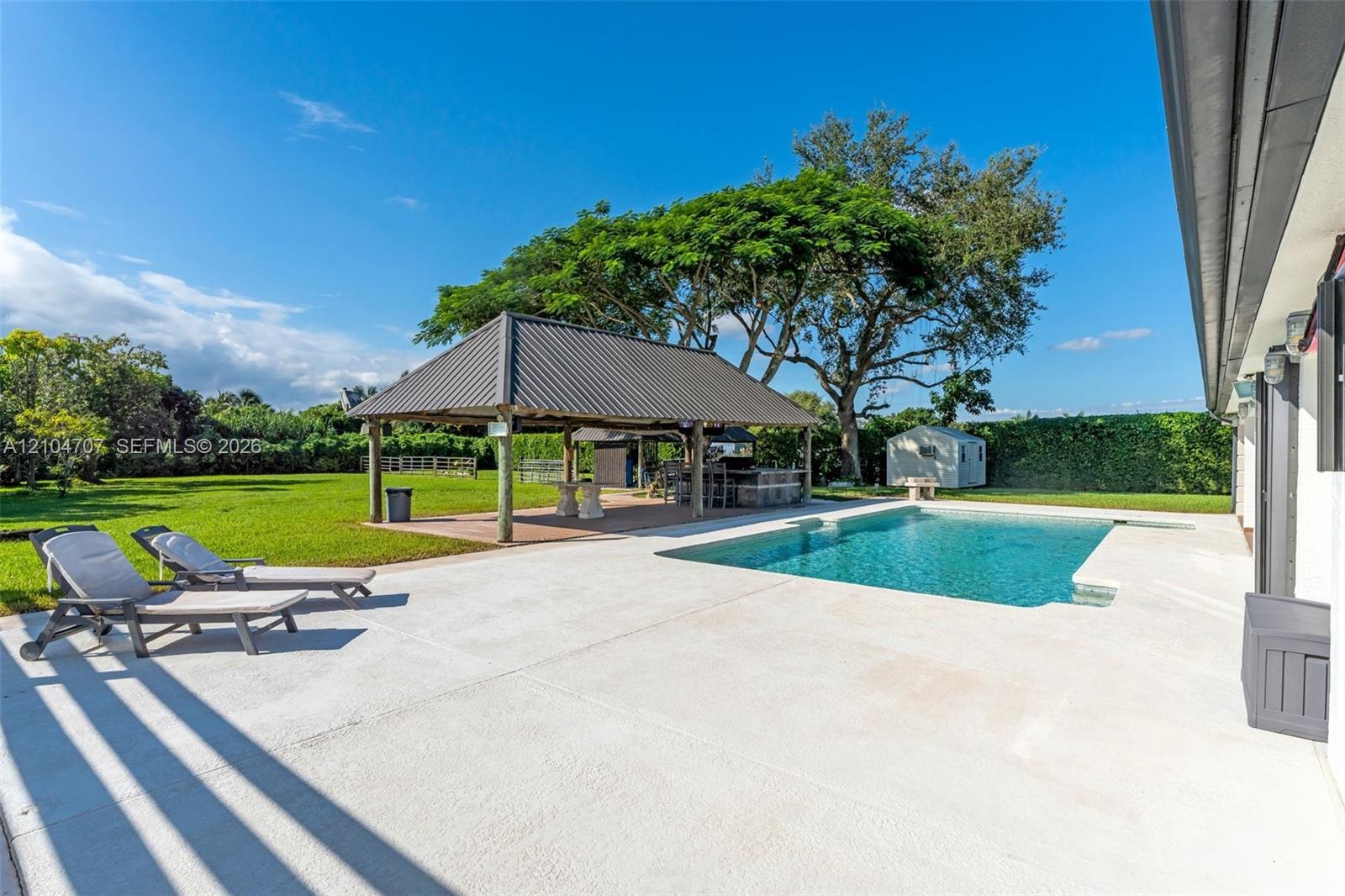 Backyard featuring a concrete pool deck, rectangular swimming pool, open-air pavilion with a metal roof, expansive grass lawn, and perimeter hedging