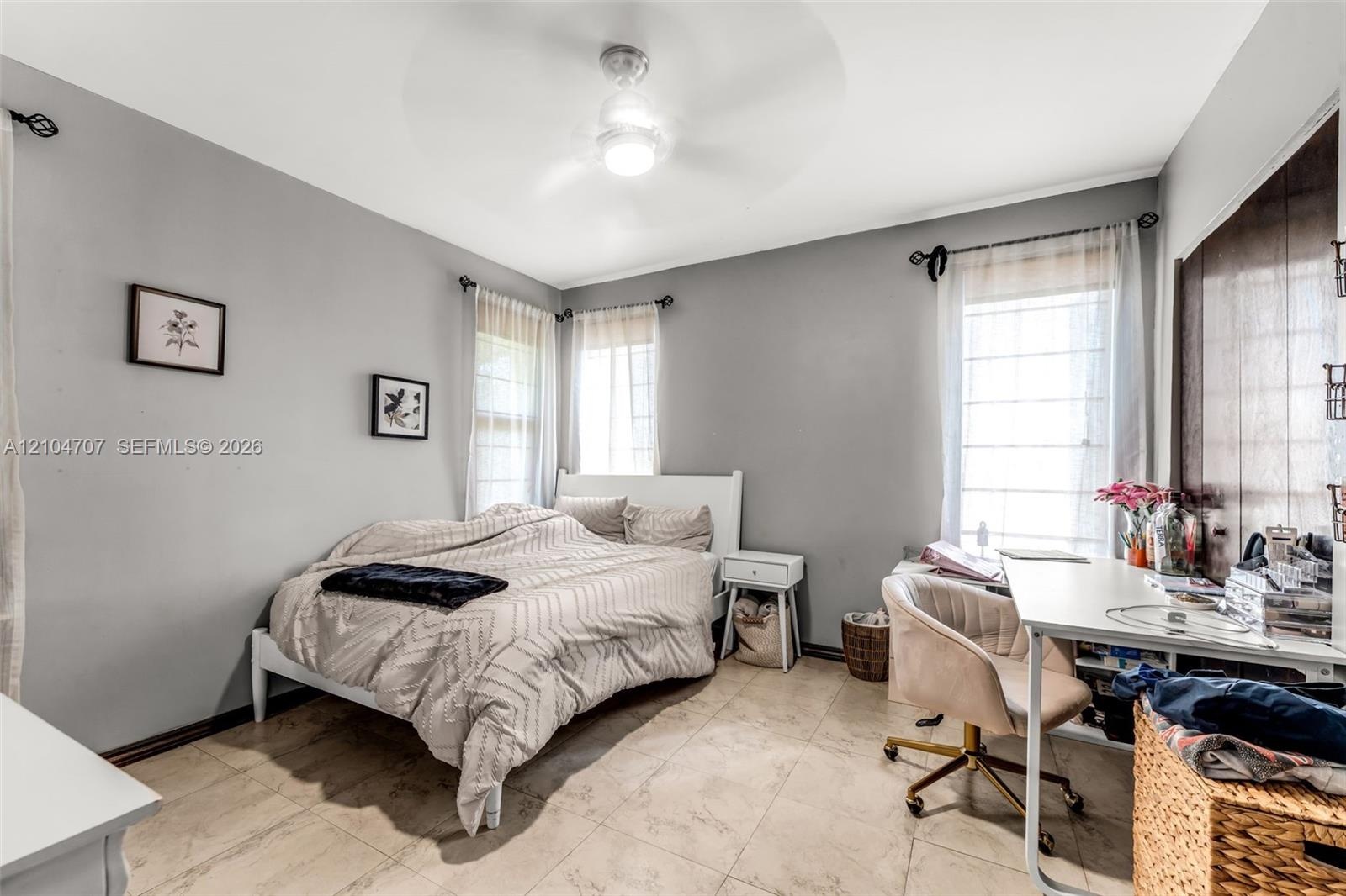 Neutral-toned interior featuring large-format tile flooring, dual-window exposure, a ceiling-mounted light fixture with fan, and bifold closet doors w