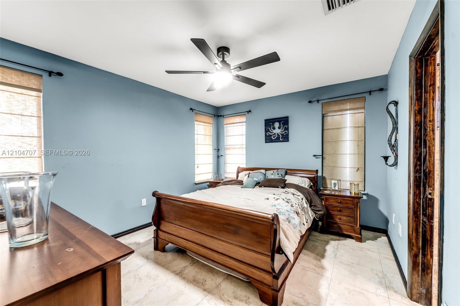 Bedroom featuring tile flooring, neutral wall color, matte black ceiling fan with integrated lighting, and three windows finished with woven-texture R