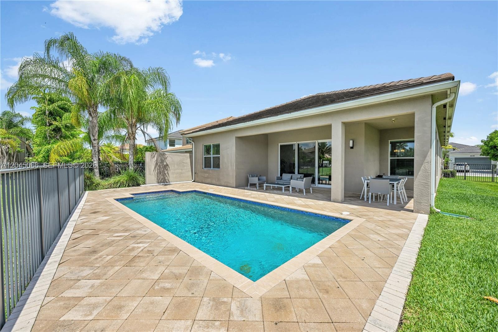 Covered patio with stone-paver flooring, rectangular swimming pool, and metal perimeter fencing overlooking a pond