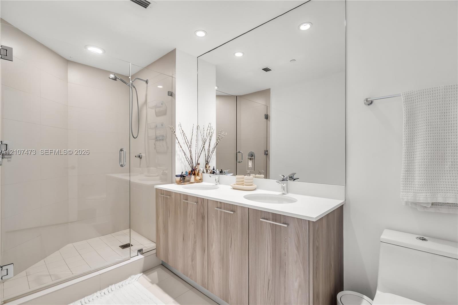 Double vanity featuring wood-finish cabinetry, solid surface countertops, dual undermount sinks, and a frameless glass-enclosed shower with tiled wall