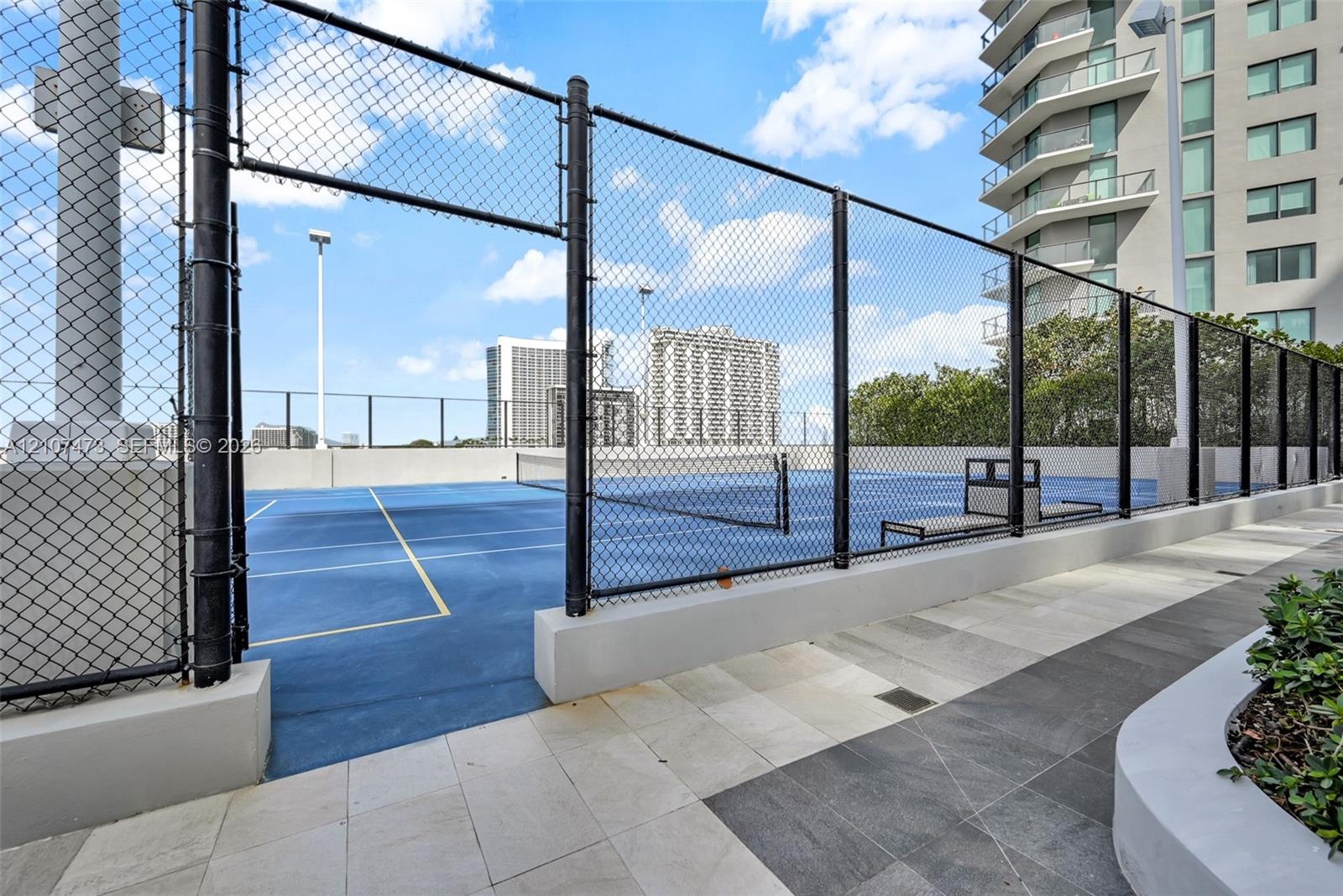 Tennis court featuring a blue hard surface, perimeter chain-link fencing, built-in court seating, and stone-tiled walking paths