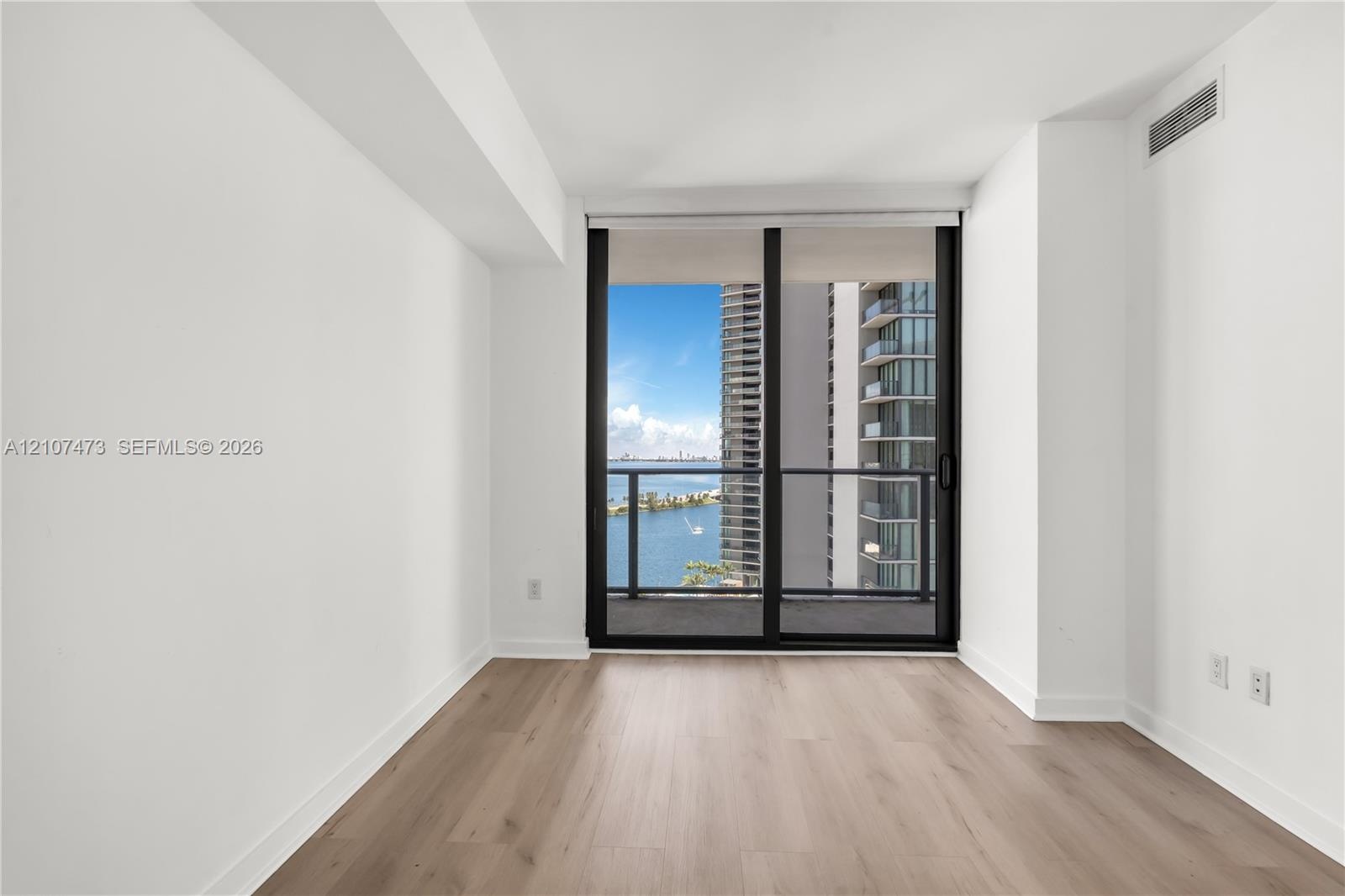 Floor-to-ceiling sliding glass doors opening to a private balcony with water and urban scenery, complemented by light wood-finish flooring