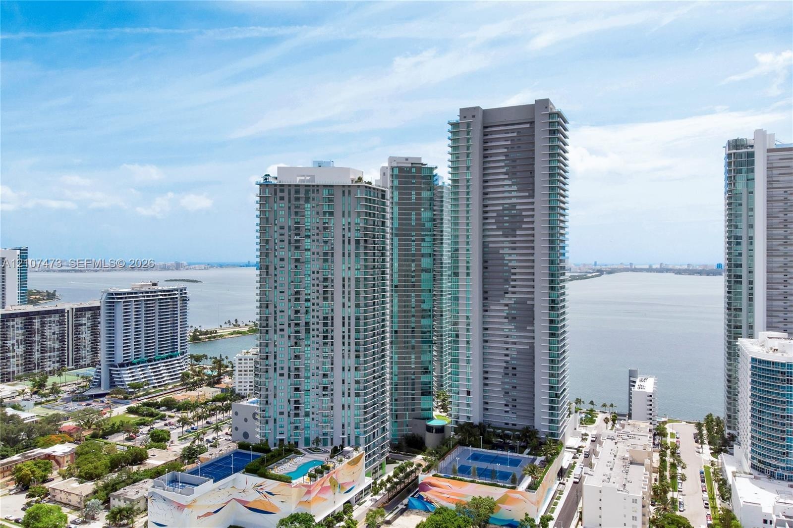 Aerial perspective of a high-rise residential complex featuring glass-facade towers, outdoor swimming pools, and tennis courts situated along the wate