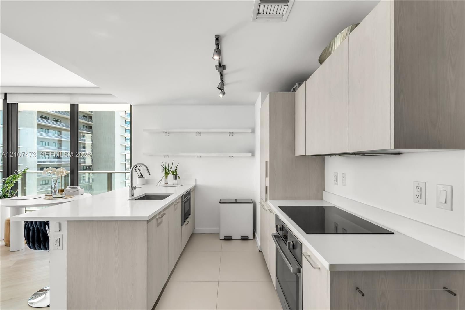 Kitchen featuring light wood-finish cabinetry, white solid-surface countertops, built-in cooktop, undermount stainless steel sink, and wall-mounted tr