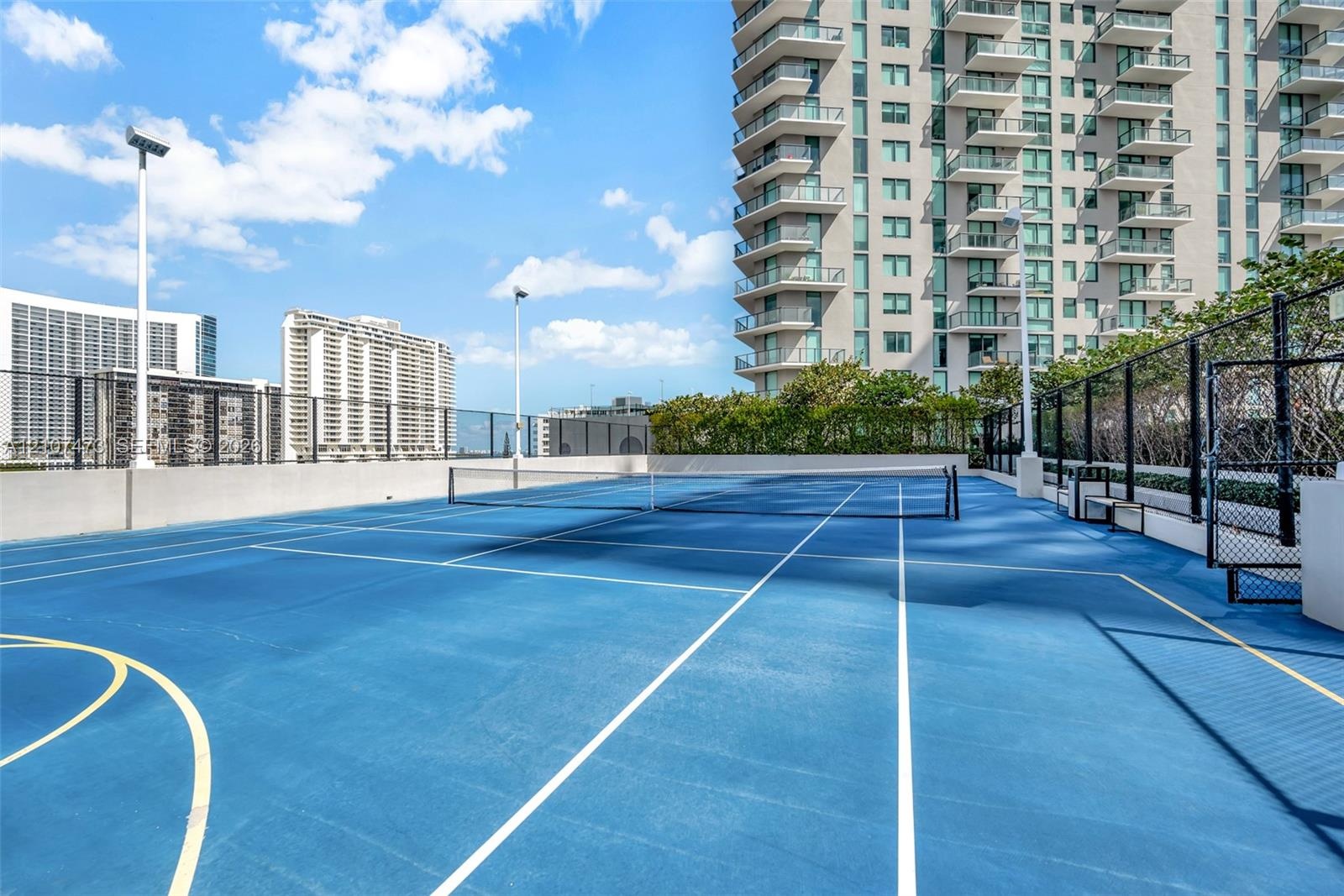 Outdoor tennis court featuring a blue hard-surface finish, perimeter chain-link fencing, court lighting, and painted lines