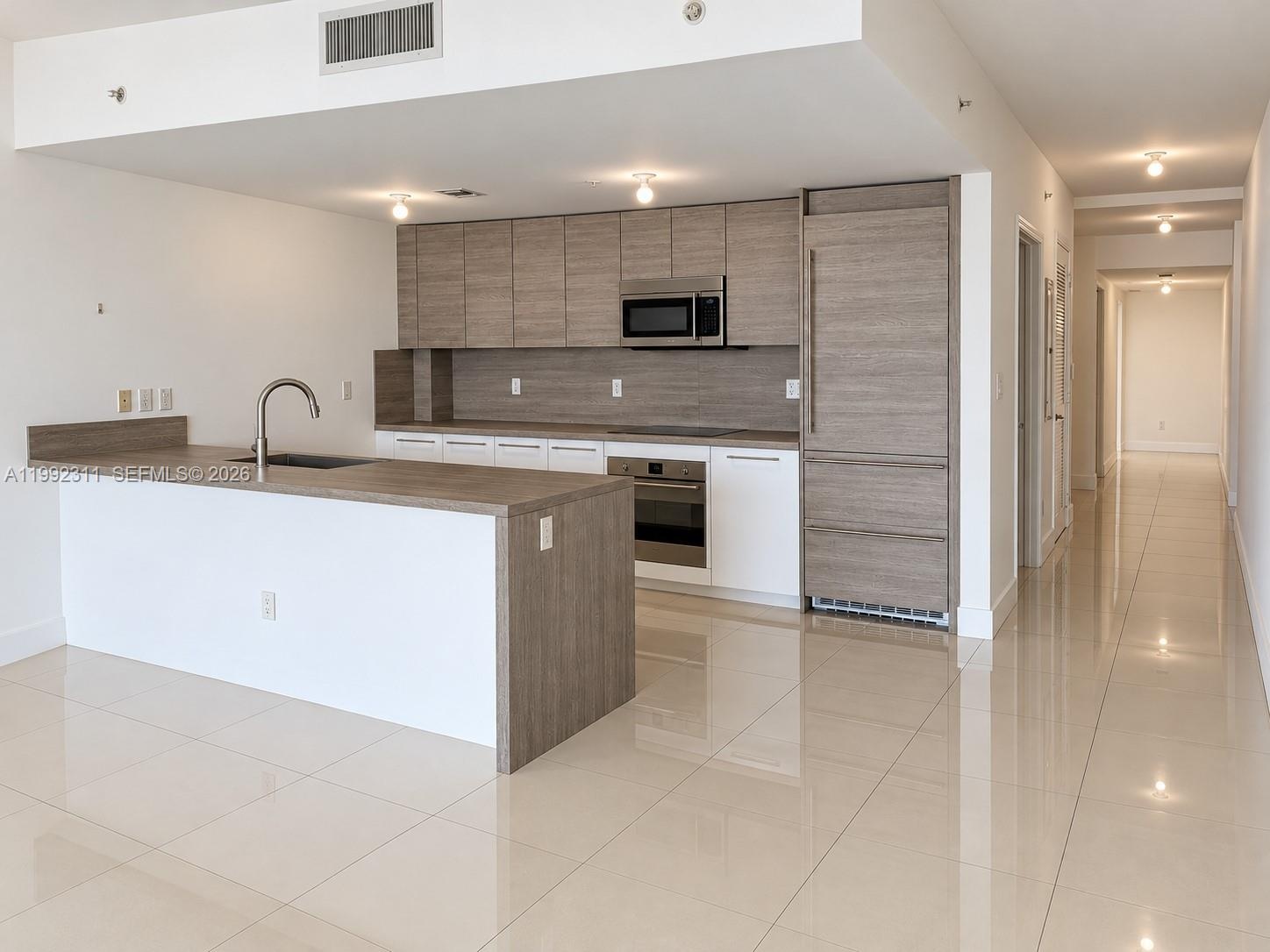 Open-concept interior featuring large-format tile flooring and a kitchen with dark wood-finish cabinetry, a peninsula counter, and integrated stainles