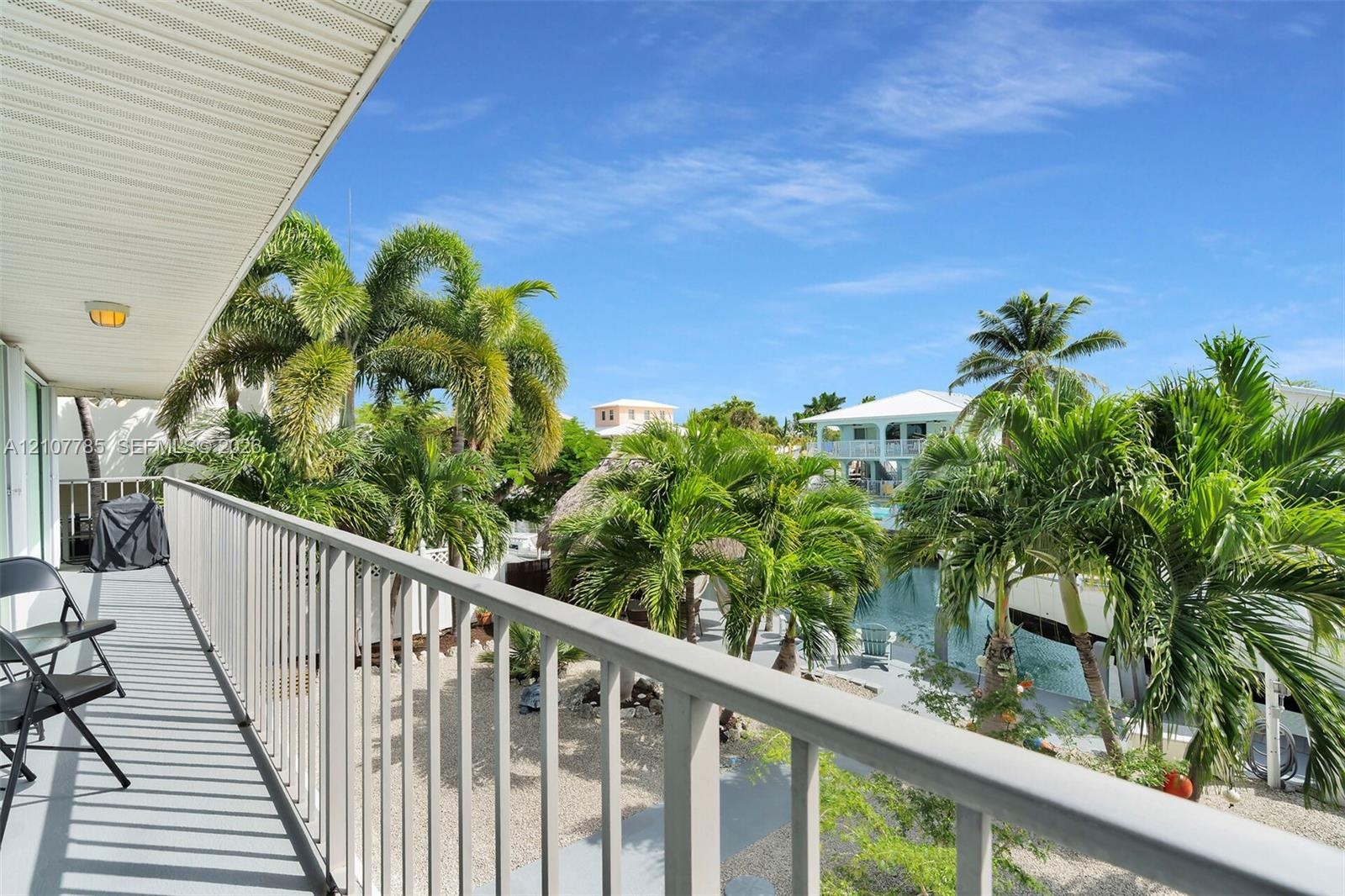 Covered balcony featuring metal railings, overhead lighting, and concrete surfacing, overlooking a canal-front exterior with palm trees and stone grou