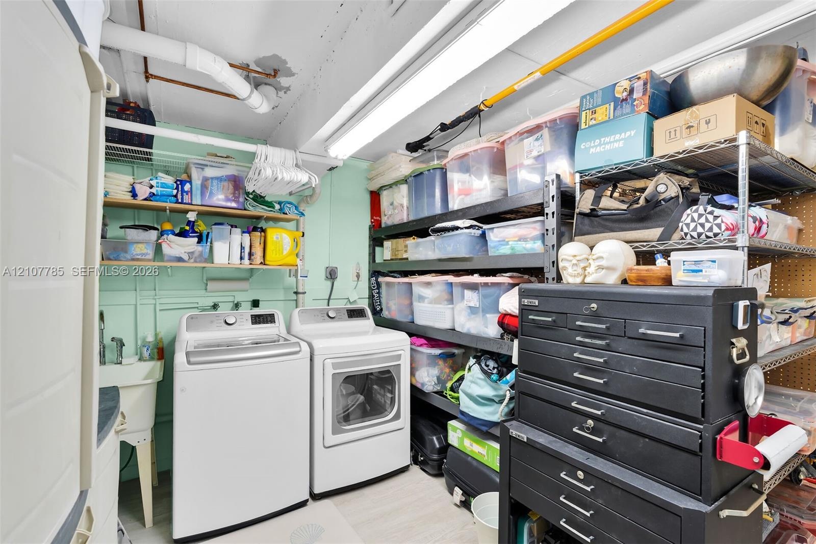 Utility room featuring a dedicated laundry station, wall-mounted utility sink, overhead fluorescent lighting, and open shelving