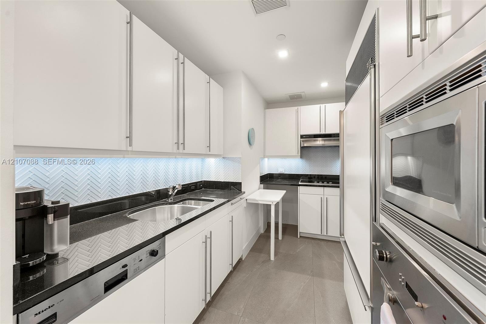 Galley kitchen featuring white cabinetry, black granite countertops, herringbone-patterned tile backsplash, stainless steel appliances