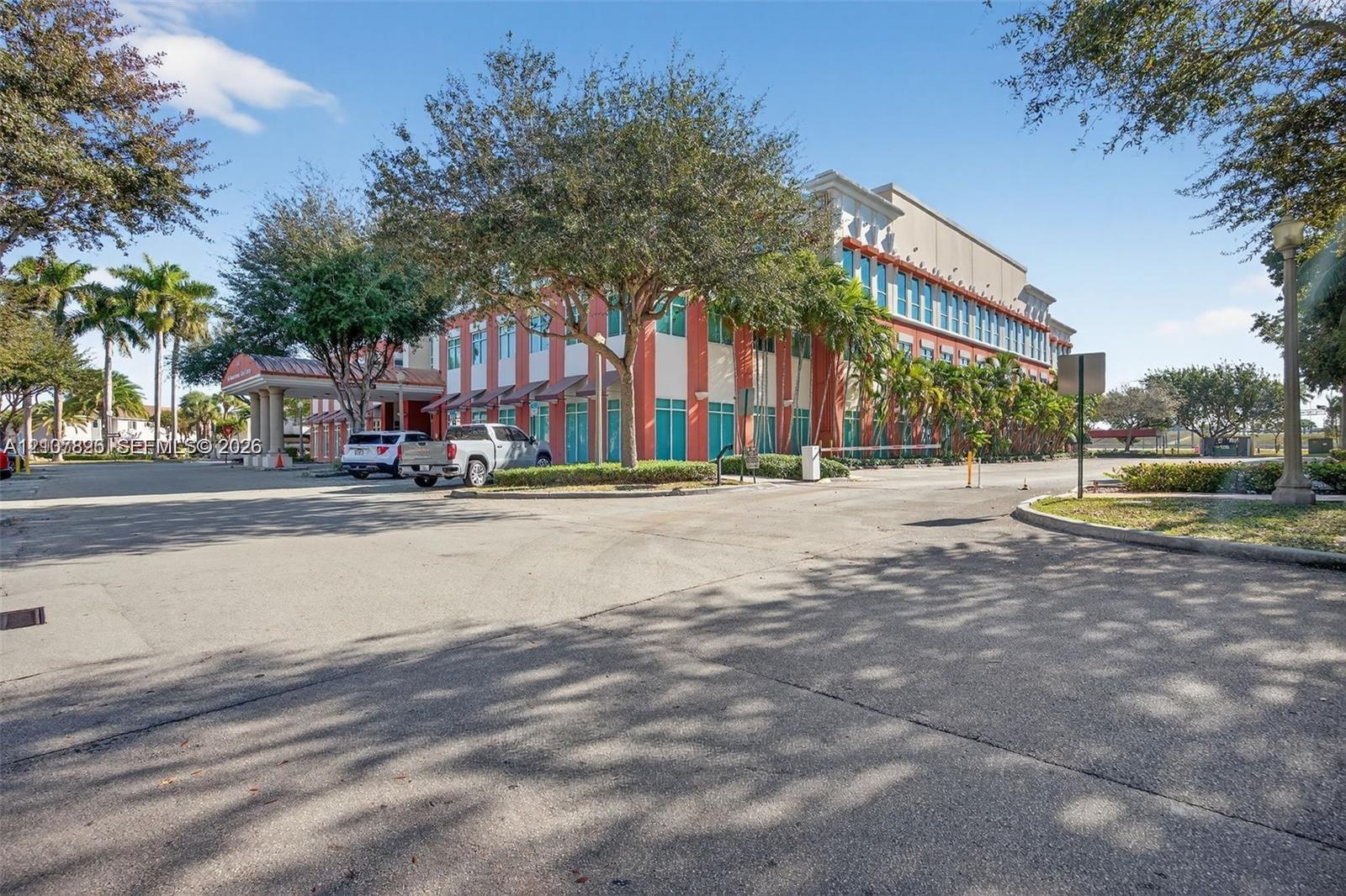 Commercial building exterior featuring a multi-story facade, covered porte-cochère, expansive paved surface parking, and mature palm and shade tree la
