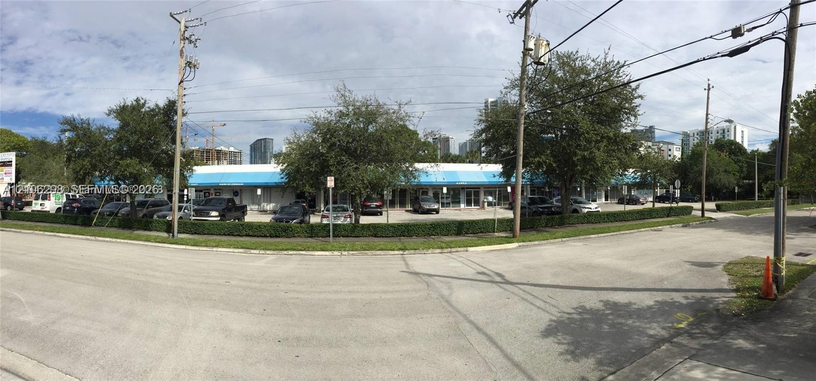 Single-story commercial plaza featuring an expansive surface parking lot, blue metal awnings, glass storefronts, and manicured perimeter hedging