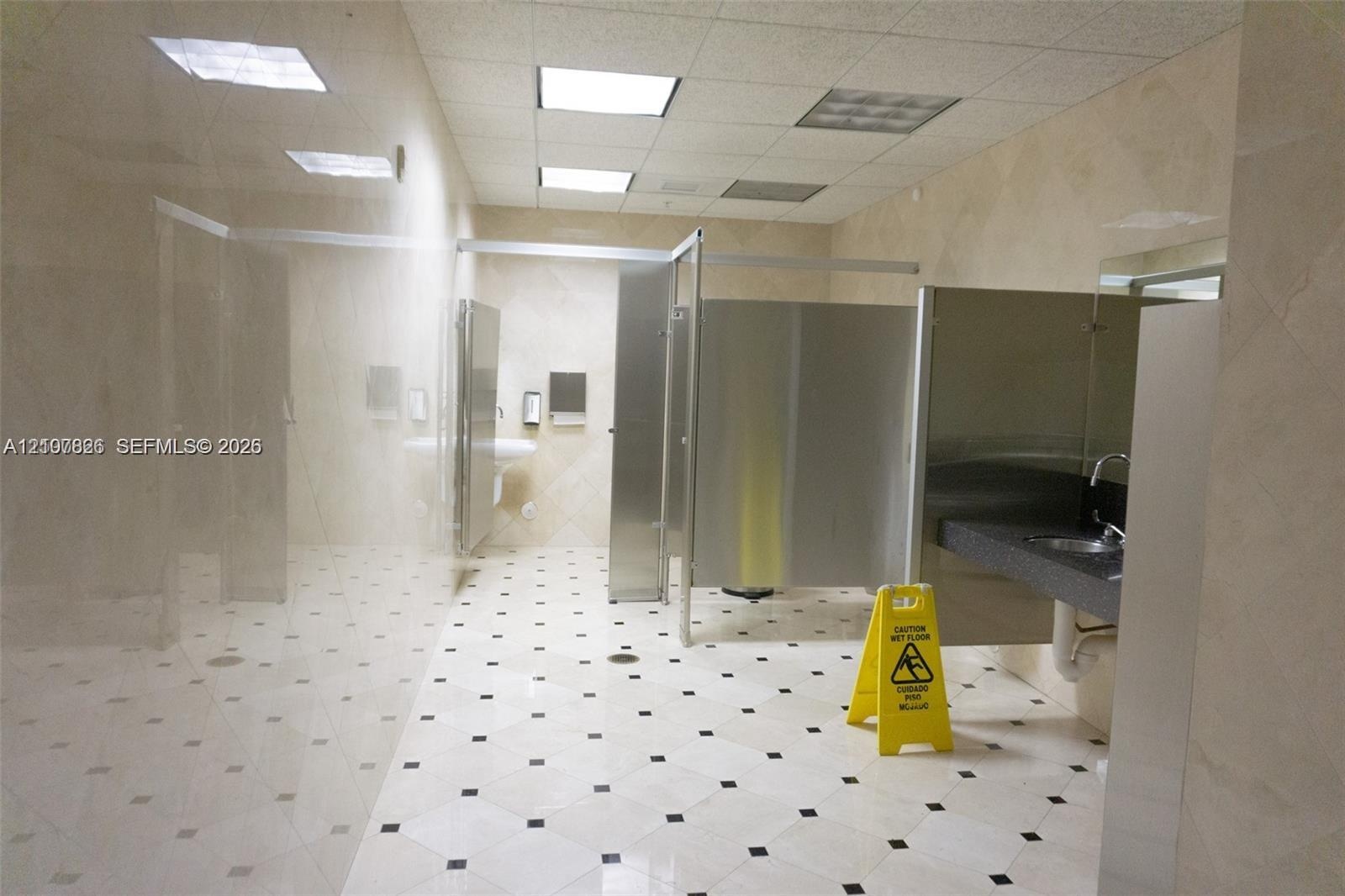 Restroom featuring floor-to-ceiling neutral wall tile, square-patterned tile flooring with dark inlays, stainless steel partitions, and a dark-toned v