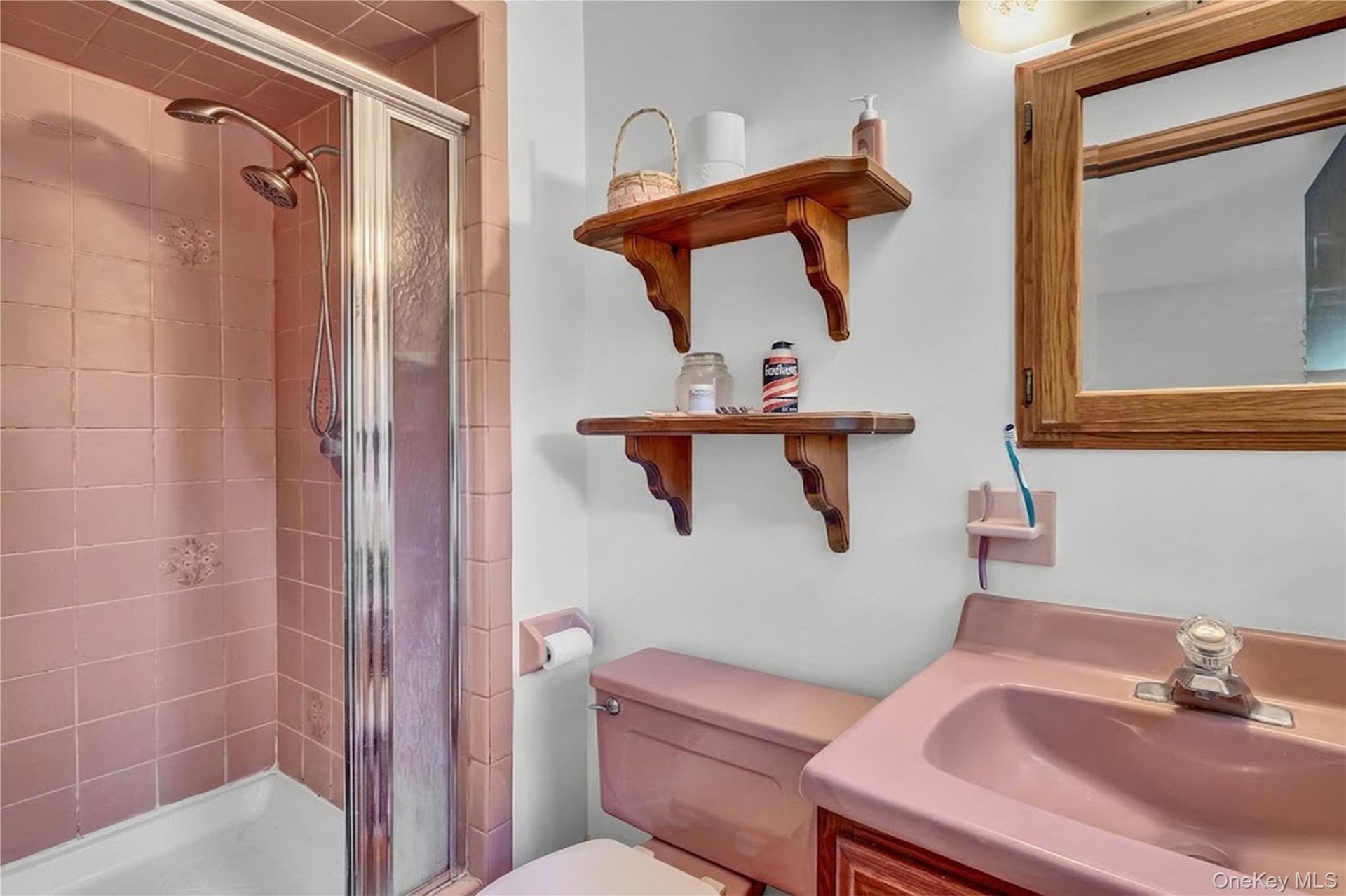 Walk-in shower with metal-framed glass enclosure, pink tile surround, integrated pink vanity with basin, wooden medicine cabinet, and wall-mounted woo