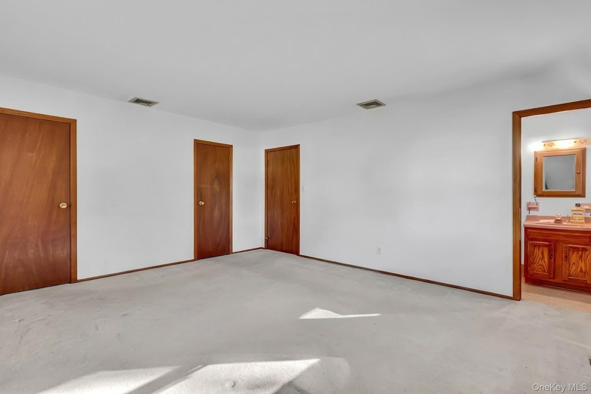 Spacious room featuring neutral carpeting, three solid wood interior doors, and an entry to an attached bathroom with a wood vanity and vanity mirror