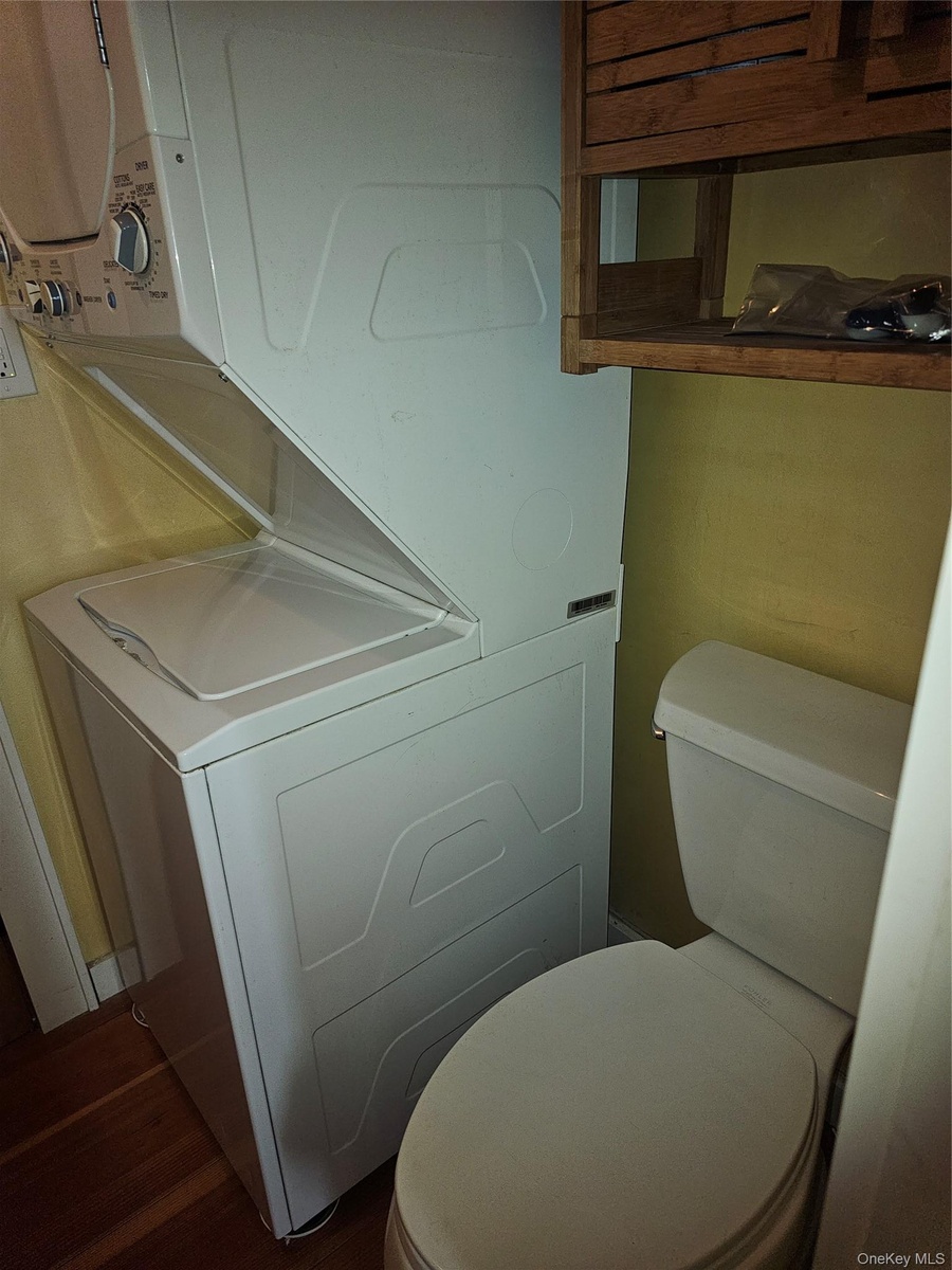Stacked white laundry center, porcelain toilet, and wall-mounted wooden shelving unit featuring wood-finish flooring