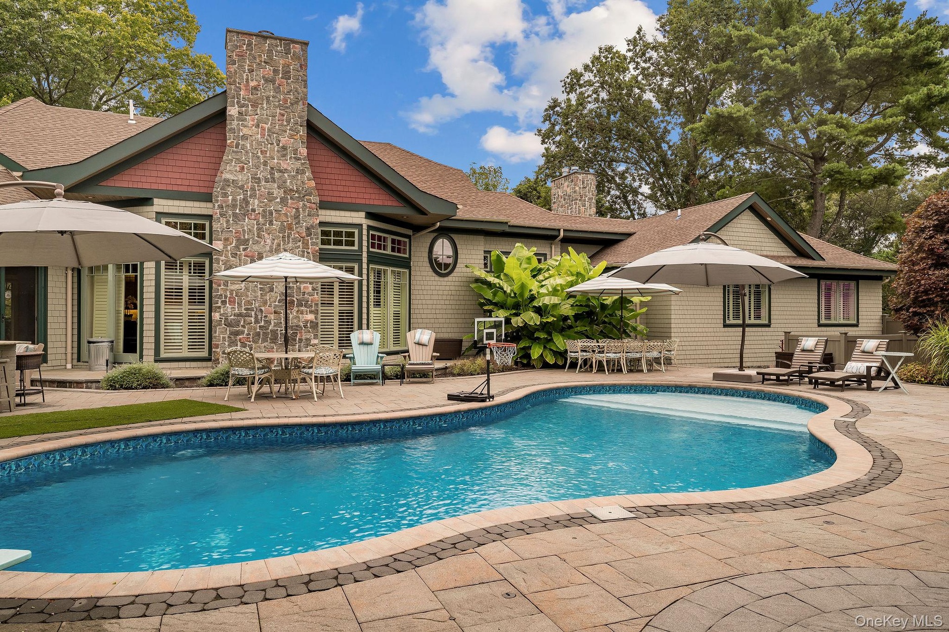 Custom-designed in-ground swimming pool with stone-tiled patio, stone exterior chimney, shingle-style siding, and extensive mature landscaping