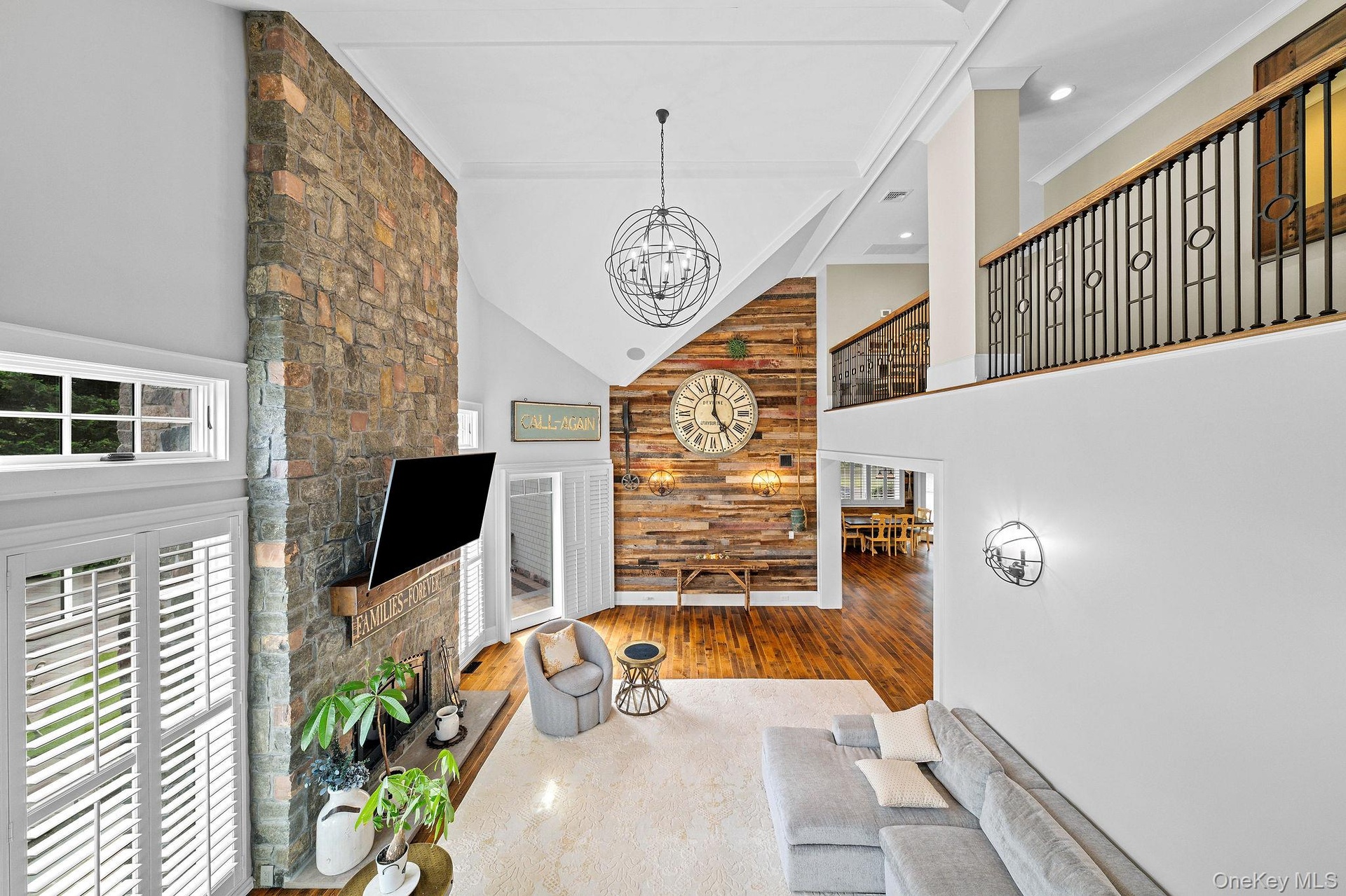 Vaulted ceilings featuring a full-height stone fireplace, reclaimed wood-finish accent wall, wood-finish flooring, and a mezzanine with wrought iron r