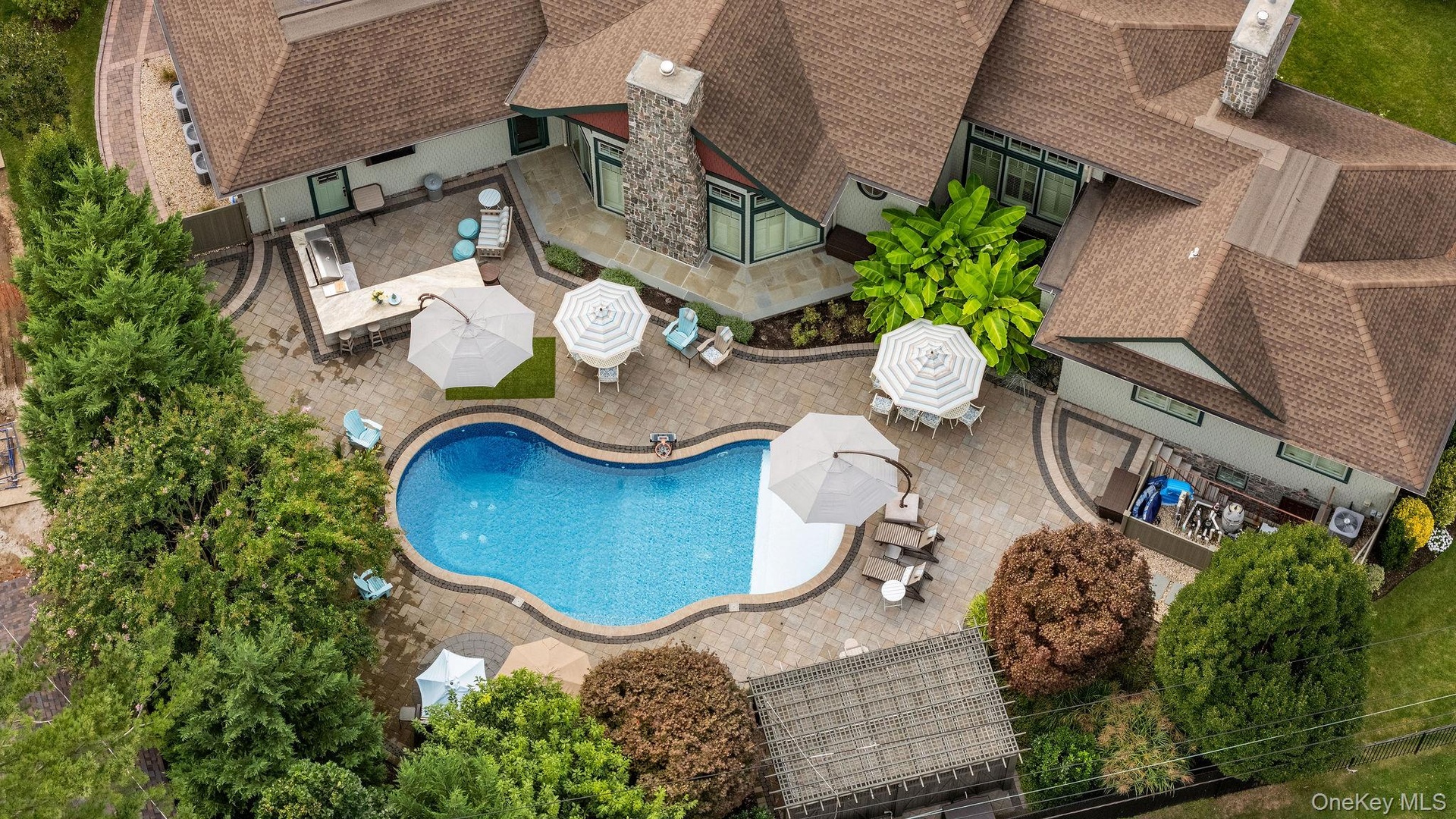 Aerial perspective of a sprawling residence featuring a custom-shaped swimming pool, extensive stone paver hardscaping, a built-in outdoor kitchen wit