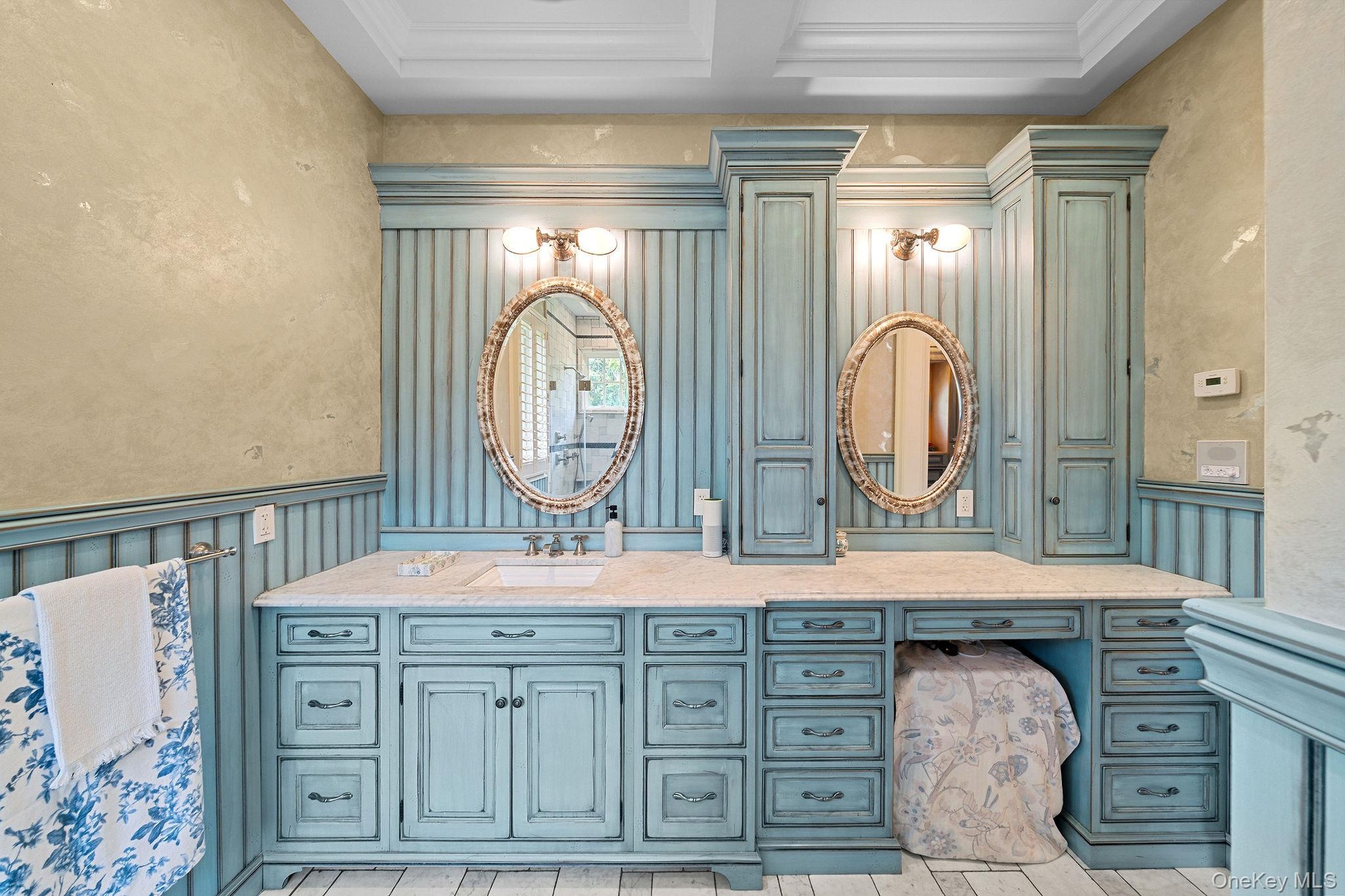 Dual-sink vanity featuring custom blue cabinetry, stone countertop, integrated floor-to-ceiling storage towers, beadboard wainscoting, and oval wall m