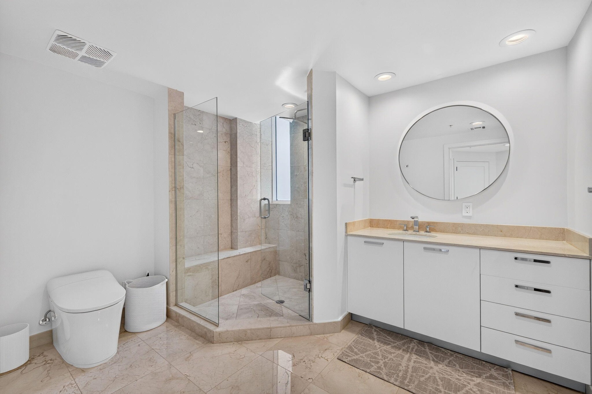 Glass-enclosed shower with a built-in bench, marble flooring and a spacious vanity featuring an integrated sink, ample storage, and a circular mirror