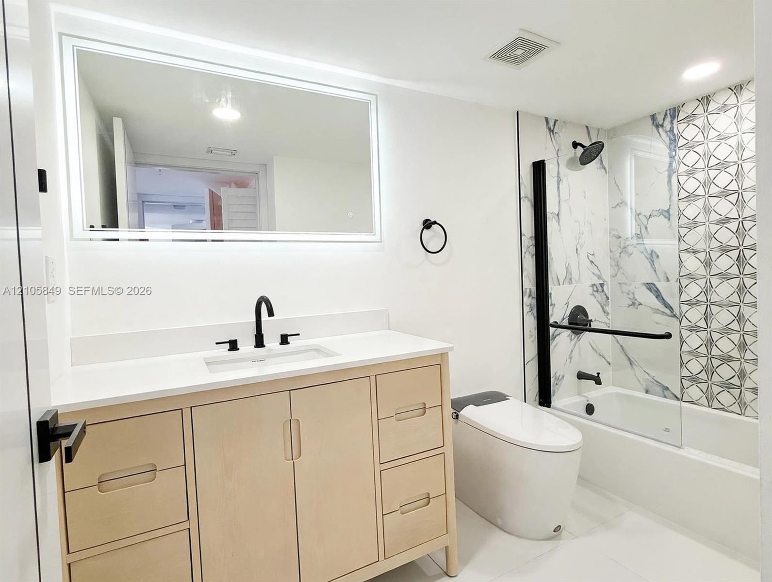 Bathroom featuring a light wood vanity with a white countertop, wall-mounted LED-backlit mirror, matte black fixtures, and a glass-enclosed tub with p