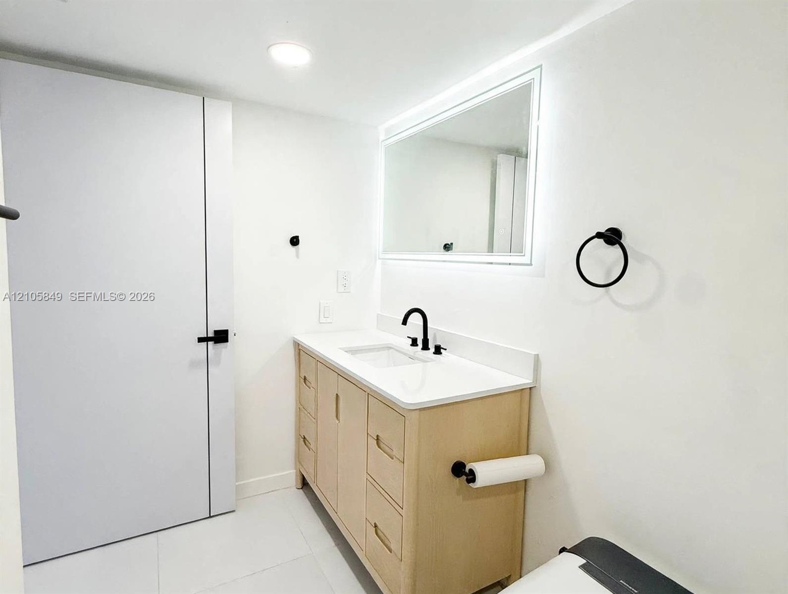 Light-finish wood vanity with a white countertop, rectangular undermount sink, integrated LED-lit mirror, and matte black fixtures