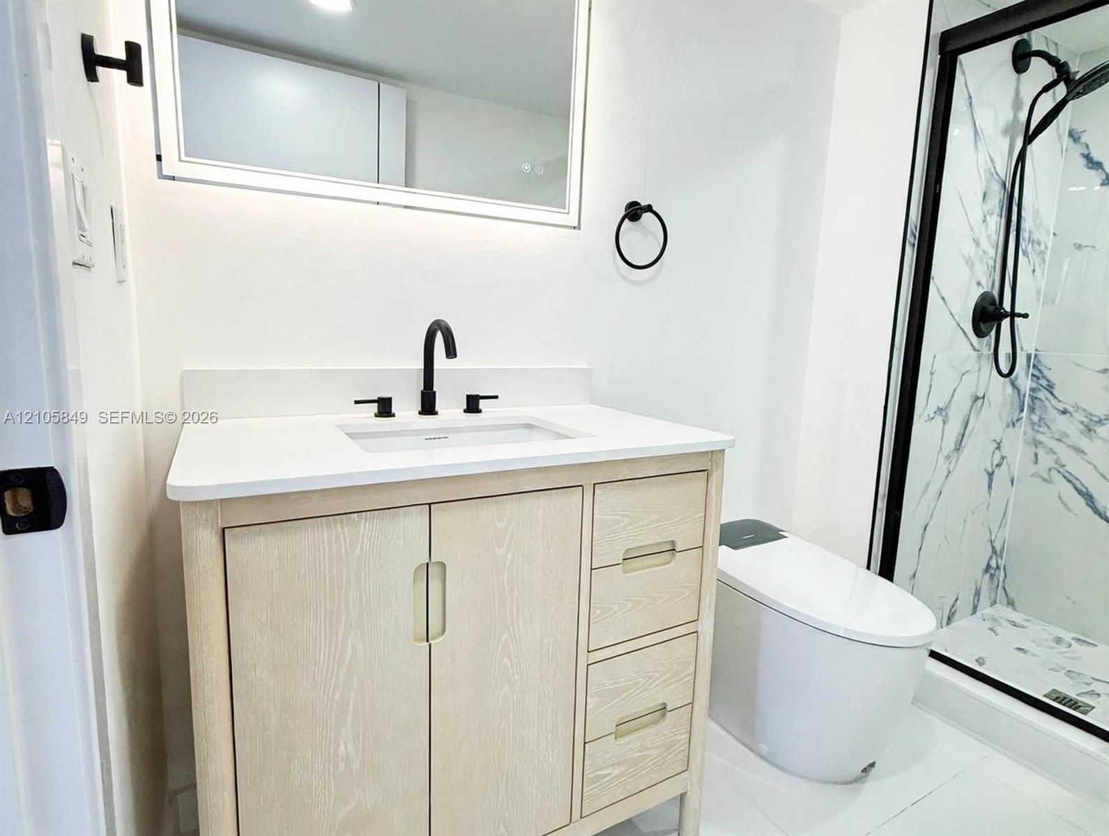Light wood-finish vanity with a white countertop, undermount sink, matte black faucet, and walk-in shower featuring patterned tile surrounds and a mat