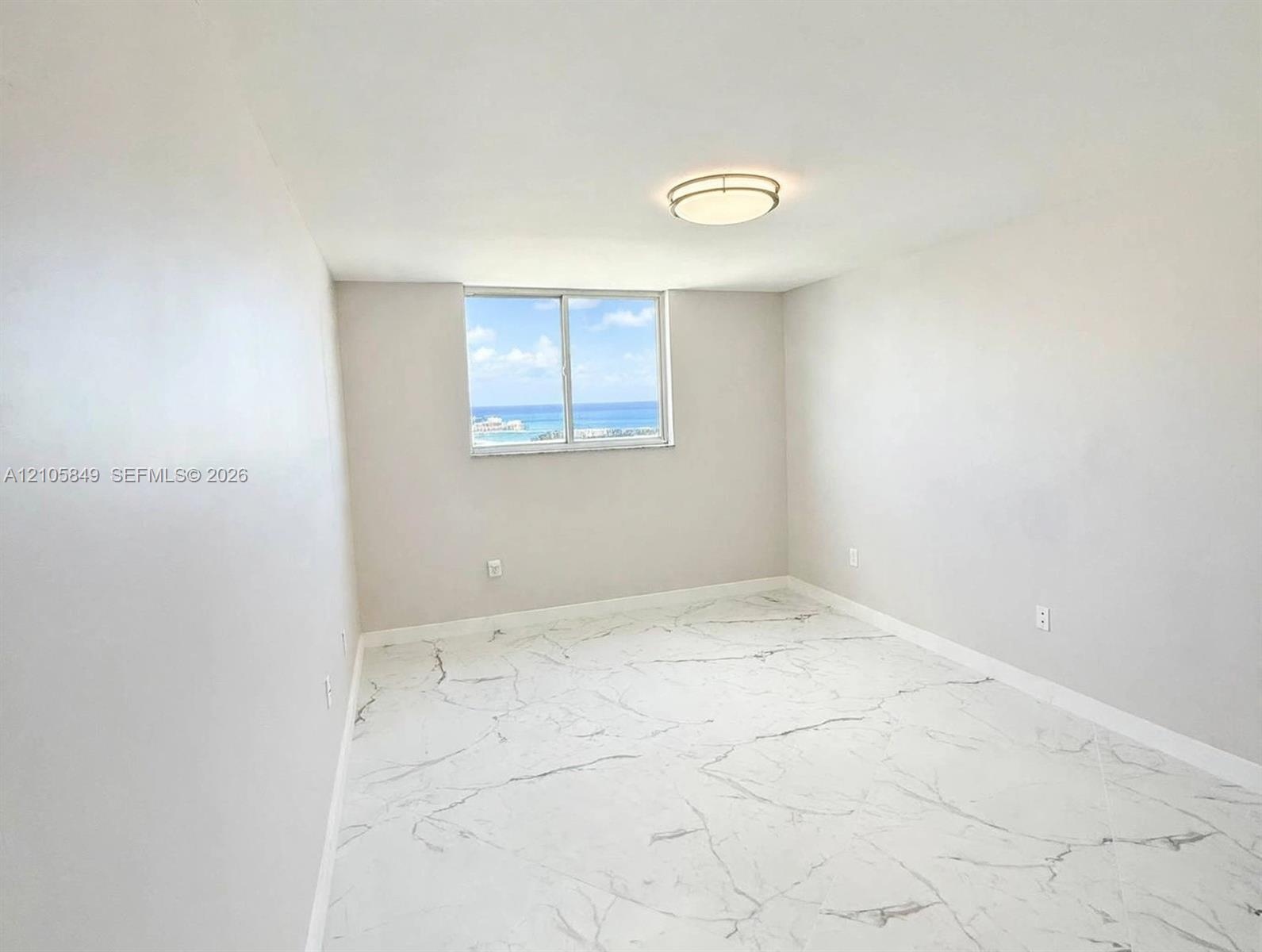 Neutral interior space featuring stone-finish flooring with veining, a flush-mount ceiling light fixture, and a window with an ocean horizon backdrop