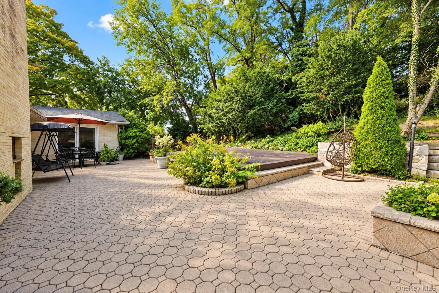 Expansive stone-paver patio featuring a built-in spa, tiered retaining walls, and mature landscaping