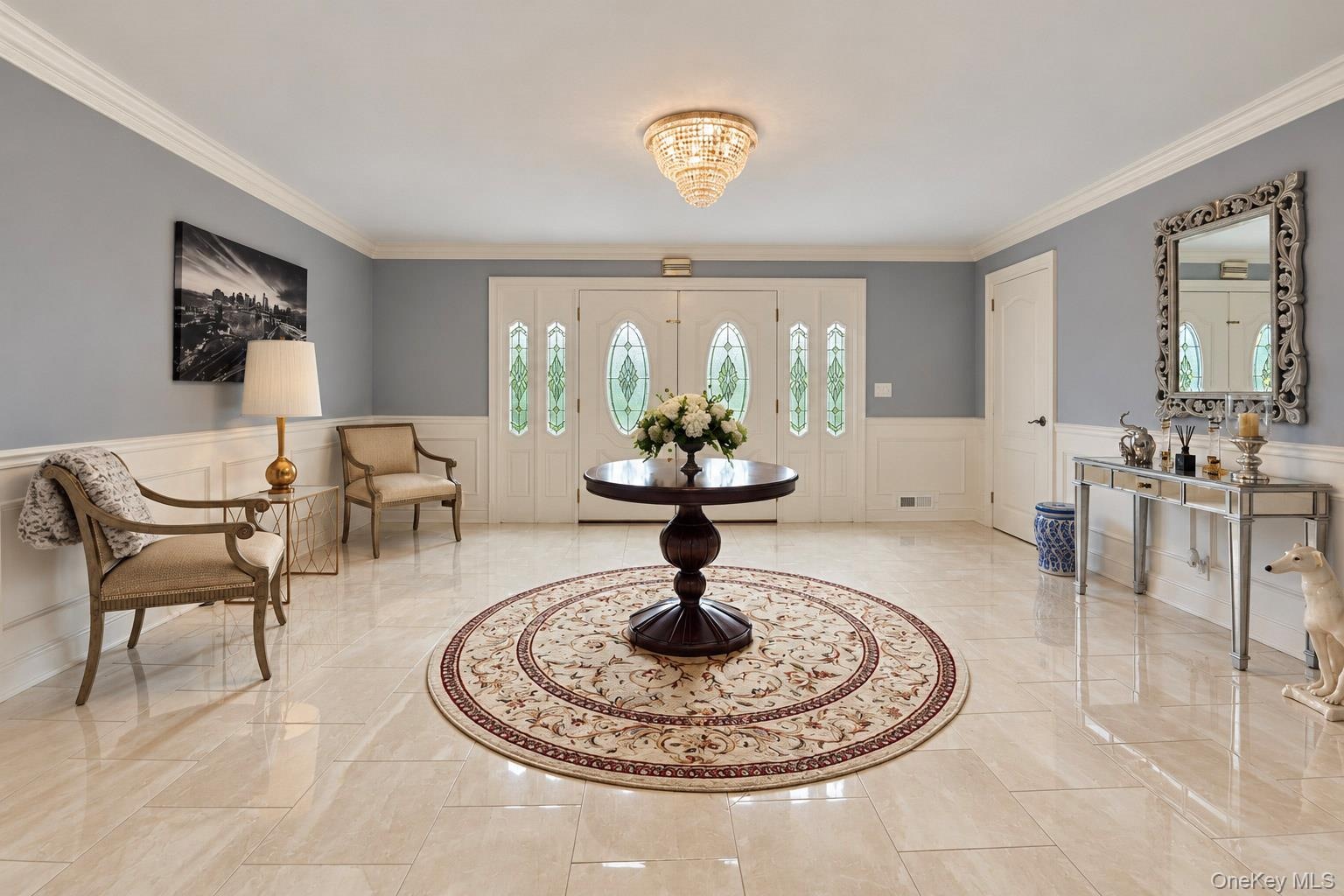 Foyer featuring large-format tile flooring, double doors with decorative glass inserts, crown molding, and wainscoting, accented by a tiered crystal-s