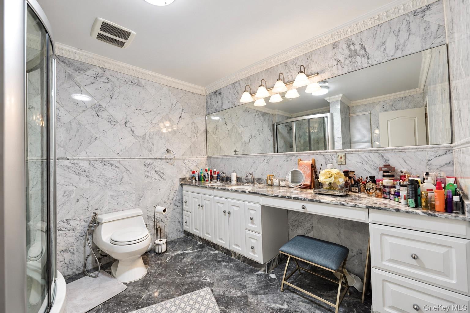 Full-bathroom featuring natural stone wall and floor tiling, expansive vanity with integrated stone countertop, crown molding, and multi-bulb wall-mou
