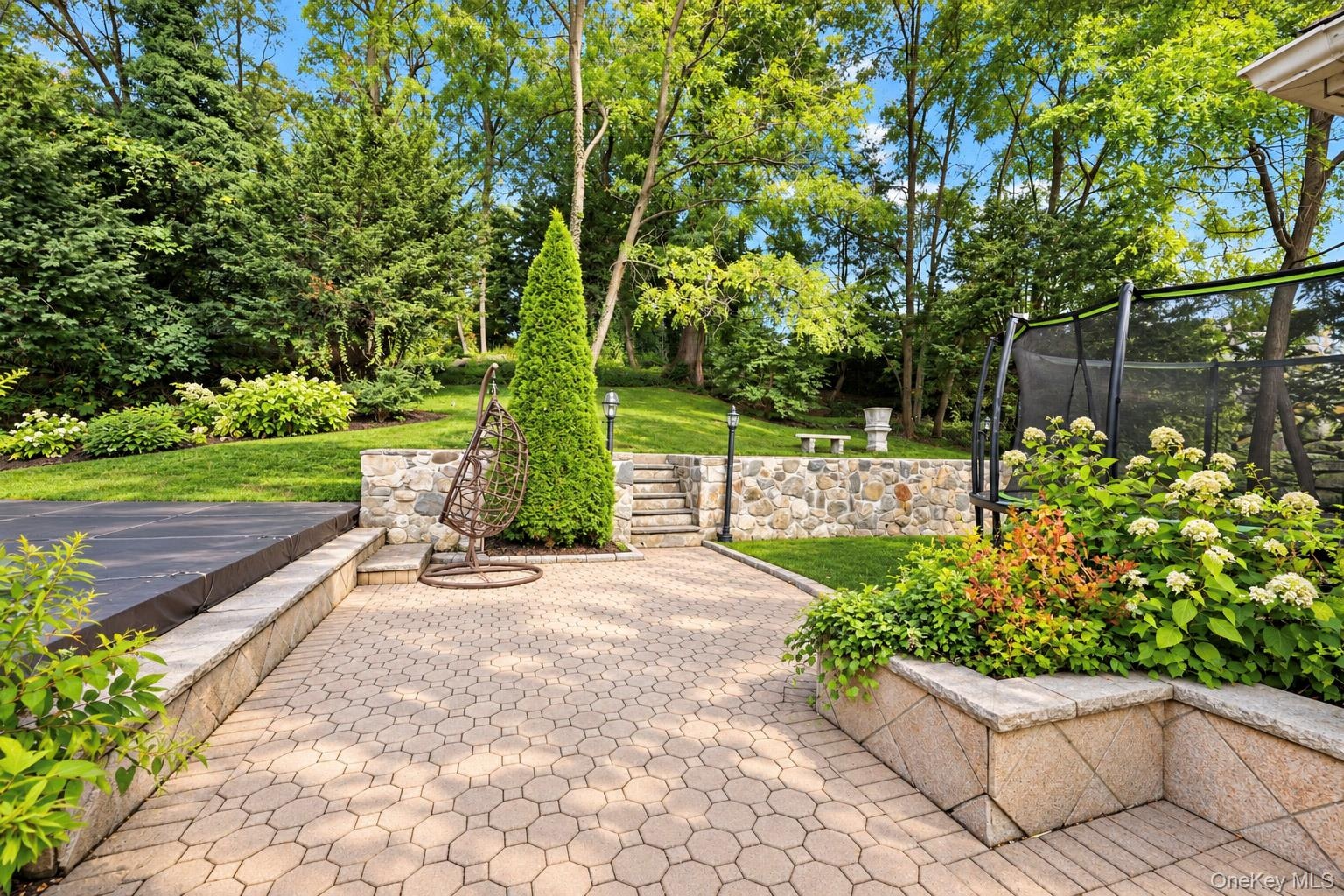 Paver patio featuring stone retaining walls, integrated stairs, tiered landscaping, and mature trees