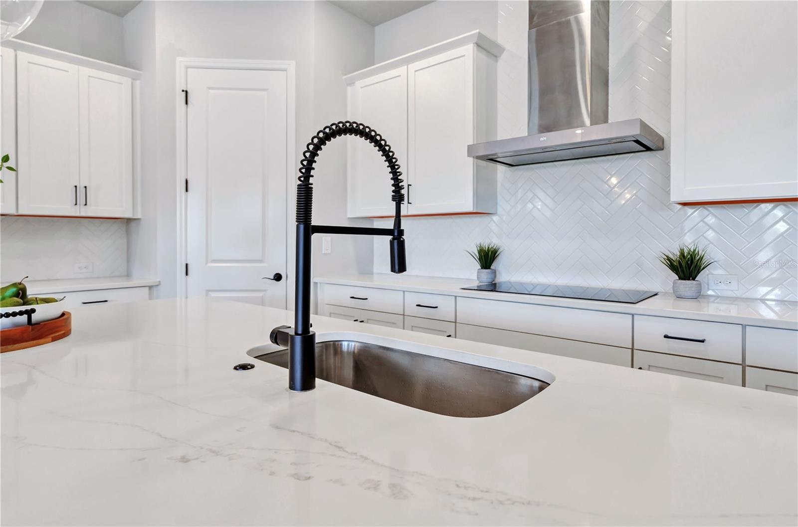 Quartz counters, undermount sink, matte black faucet, and herringbone tile backsplash with a view into the home.