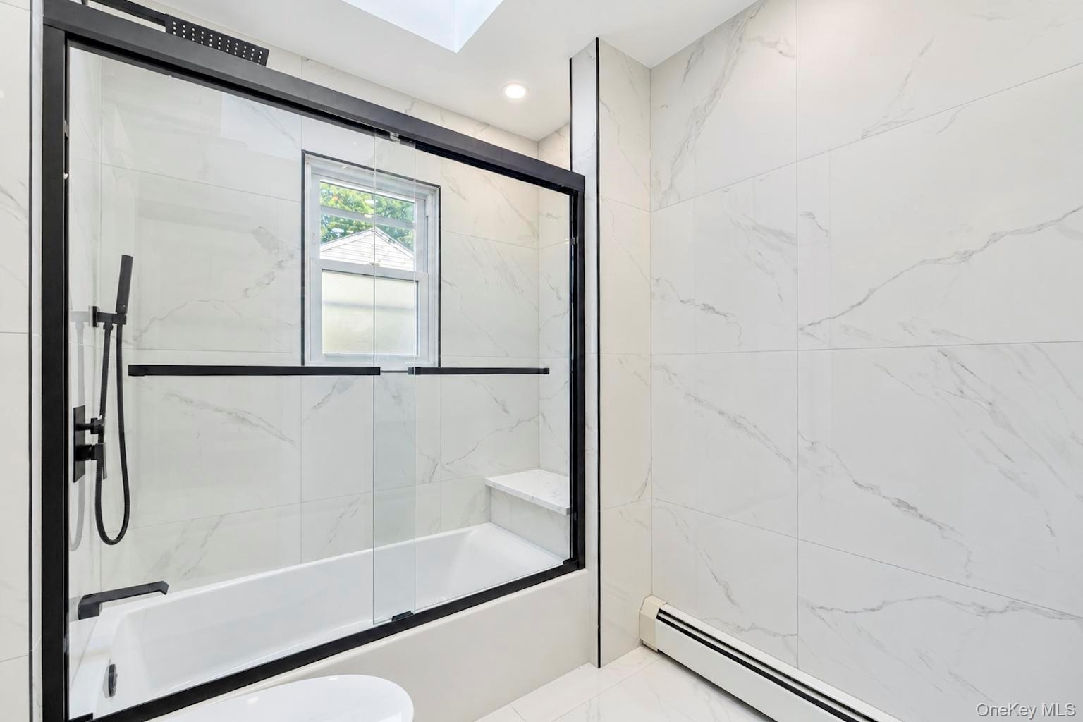 Full-height stone-finish wall tile, bathtub with integrated bench seating, black-framed glass sliding shower enclosure, matte black rainfall shower he