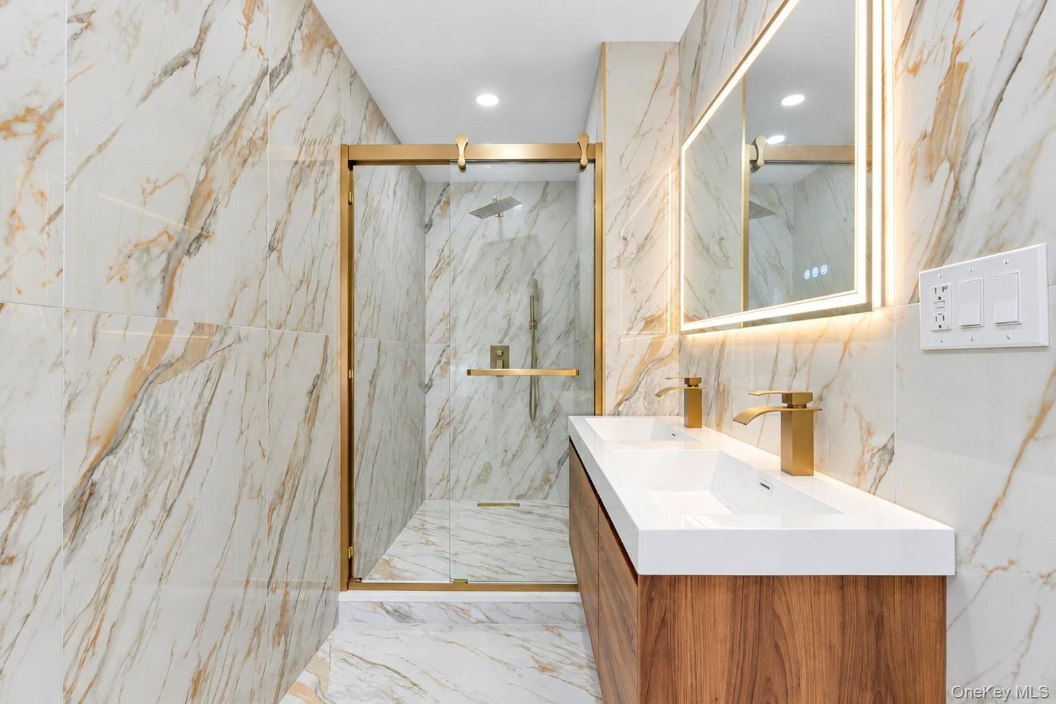 Dual vanity with integrated sinks, wall-to-wall large-format porcelain tile, walk-in shower with sliding glass door, brass-finish fixtures, and integr