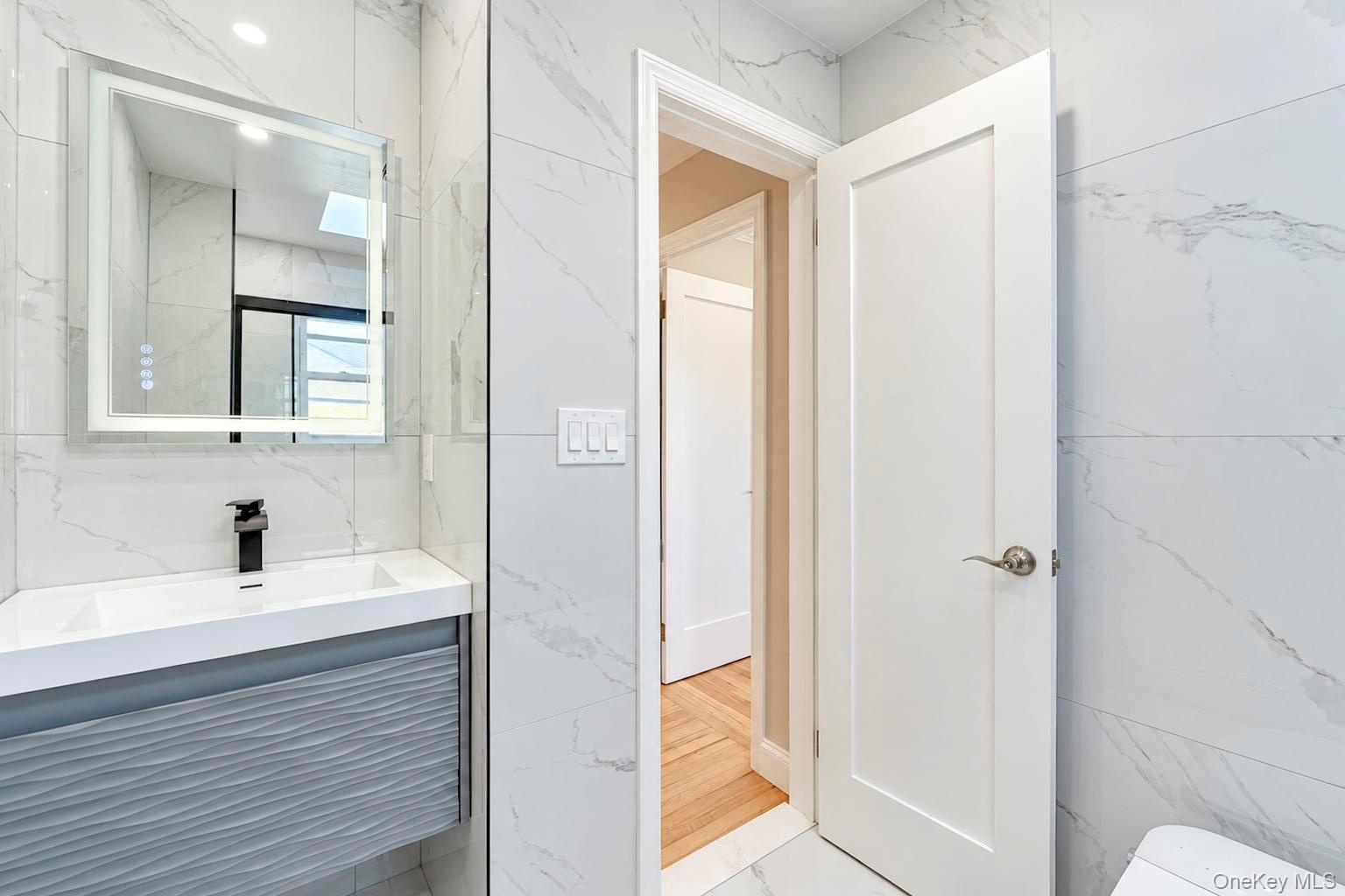 Floating vanity with a textured grey facade, integrated rectangular sink, wall-mounted matte black faucet, illuminated vanity mirror, and large-format