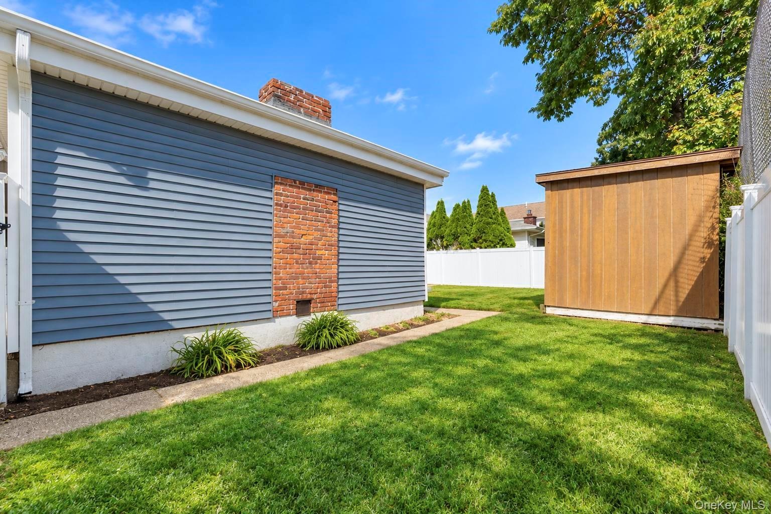 Exterior side yard featuring blue lap siding, exposed brick masonry chimney detail, manicured lawn, detached storage structure, and vinyl privacy fenc