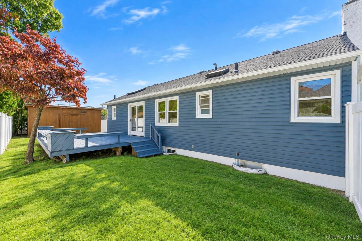 Exterior featuring blue horizontal siding, raised wooden deck, manicured lawn, white vinyl fencing, and a detached storage structure