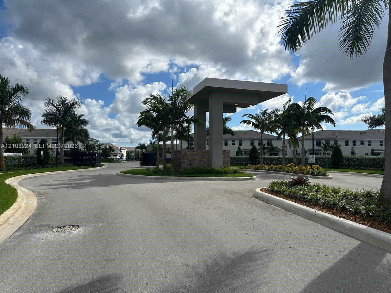 Entryway featuring a modern geometric gatehouse, stone-veneer pillar accents, asphalt paving, and established palm landscaping