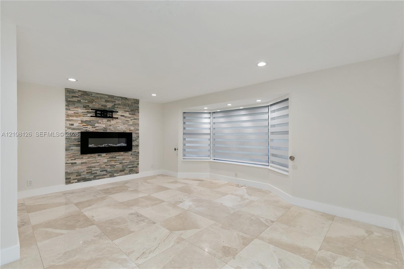 Stone-accented fireplace with a wall-mounted fire feature, large bay window, and light-toned tile flooring
