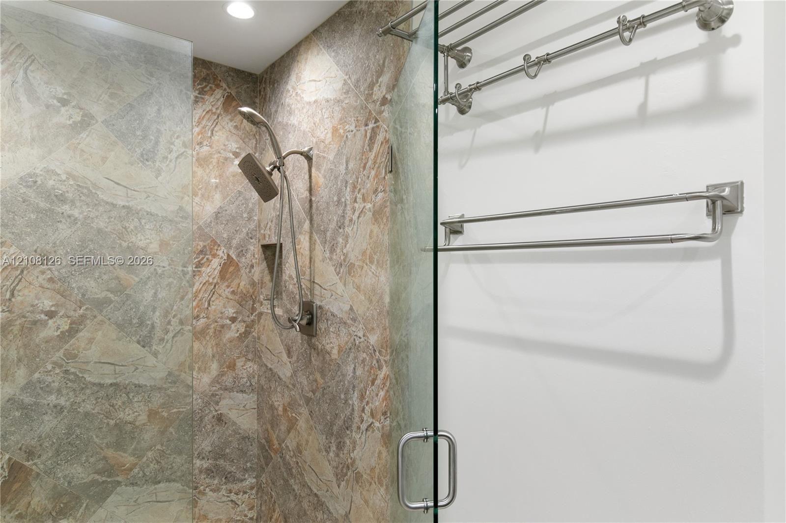 Walk-in shower featuring large-format tile surround, adjustable handheld shower fixture, built-in recessed storage niche, frameless glass door, and br