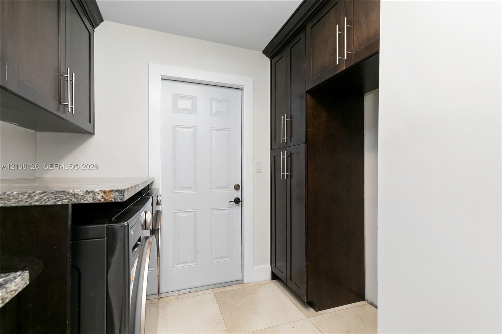 Dark-toned cabinetry featuring stainless steel hardware, stone-finish countertops, tile flooring, and a paneled exterior door