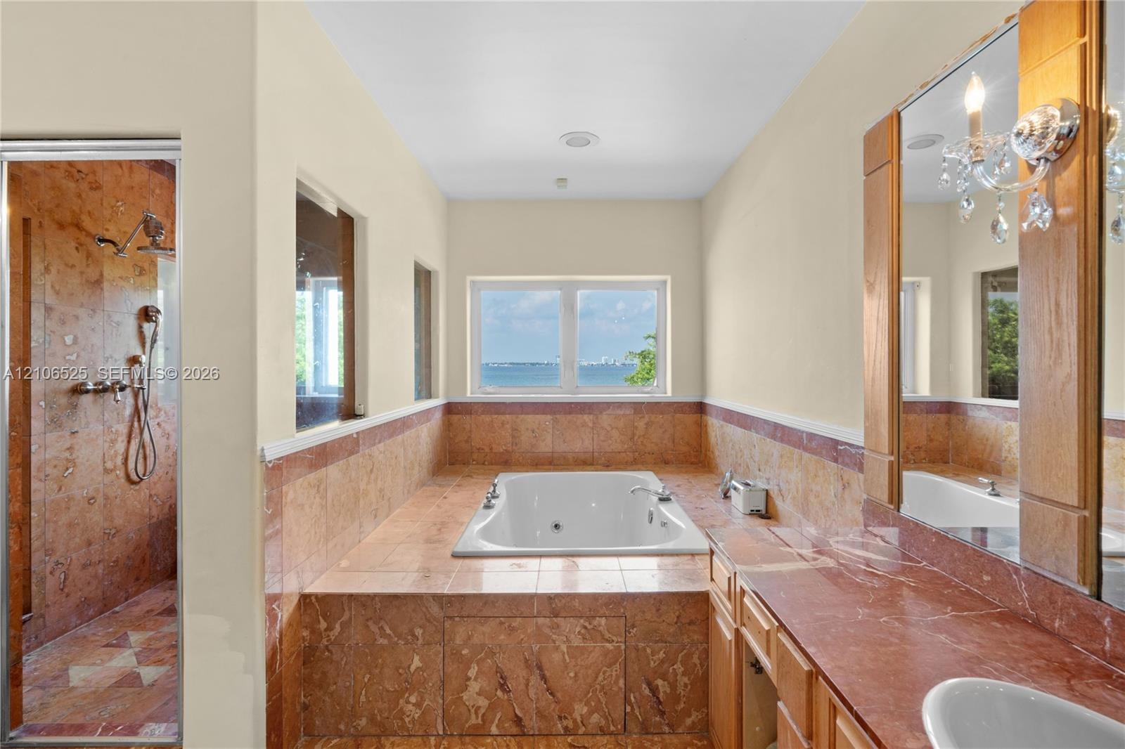 Bathroom featuring a built-in soaking tub, natural stone tile surround, walk-in shower with stone tile walls, stone-topped vanity, and wall-mounted li