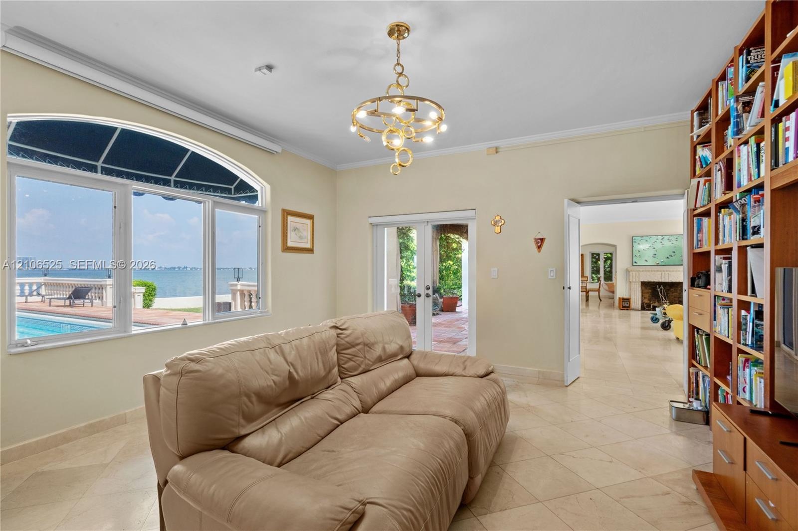 Large arched window with water and pool expanse, built-in floor-to-ceiling wood shelving, gold-tone contemporary chandelier, and light-colored stone f